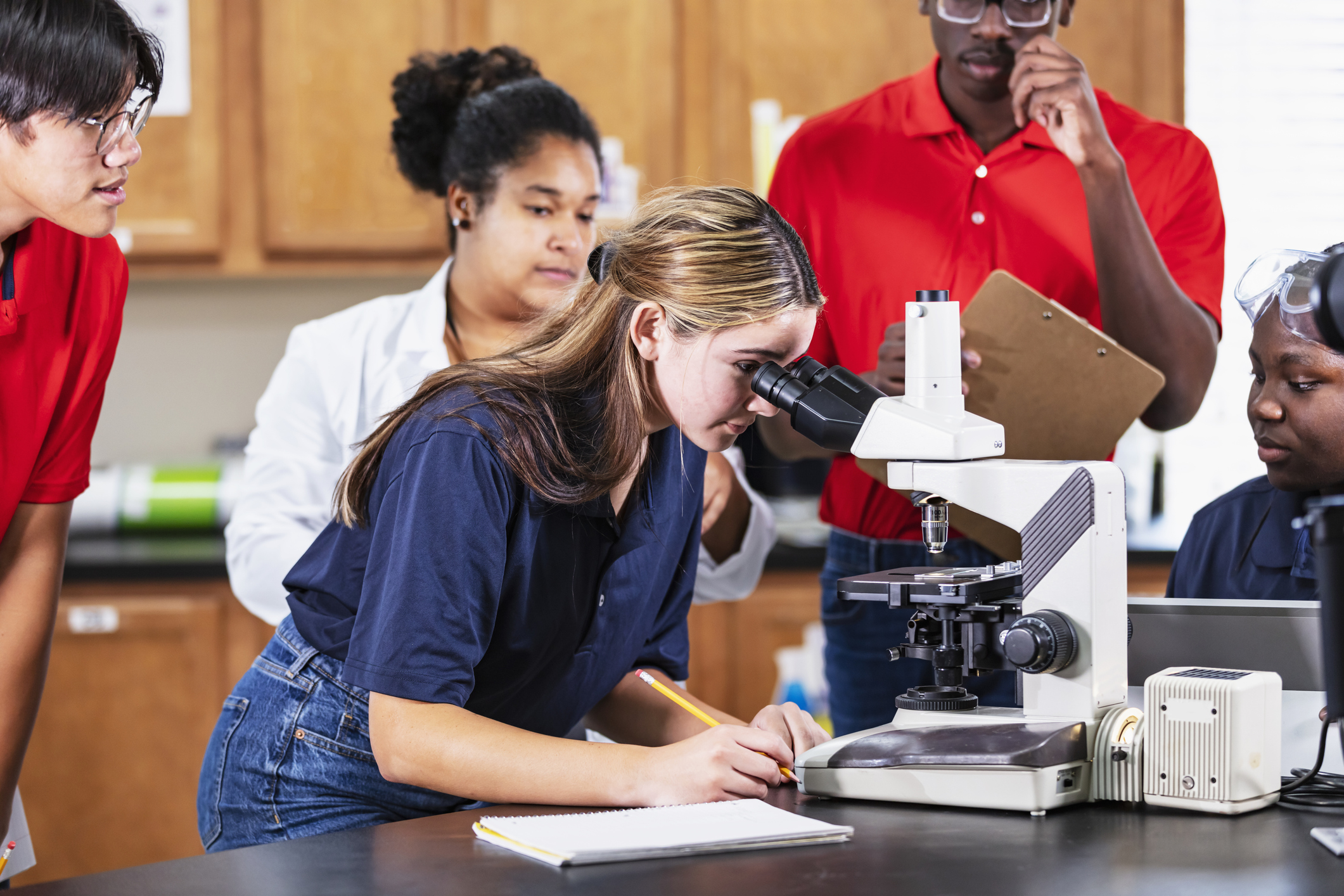 STEM student looking through microscope and taking notes with other students and teacher behind her